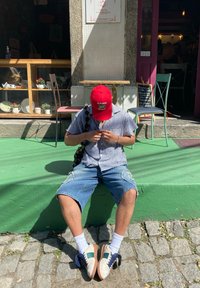 Indivíduo sentado num degrau verde, usando um boné vermelho, camisa xadrez e calções de denim rasgados. Os sapatos são bege com detalhes em azul e verde.