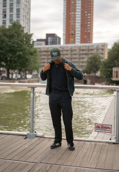 Sudadera con cremallera de color verde oscuro sobre una camiseta negra, pantalones negros y zapatillas Adidas negras. Lleva una gorra verde con texto blanco. Madero junto al agua.