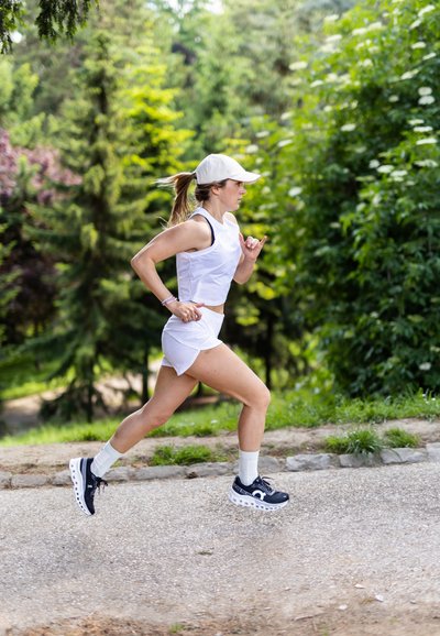 Mujer corriendo en un área boscosa, vistiendo una camiseta sin mangas blanca, pantalones cortos blancos y zapatillas deportivas azul marino con suelas blancas y cordones negros.