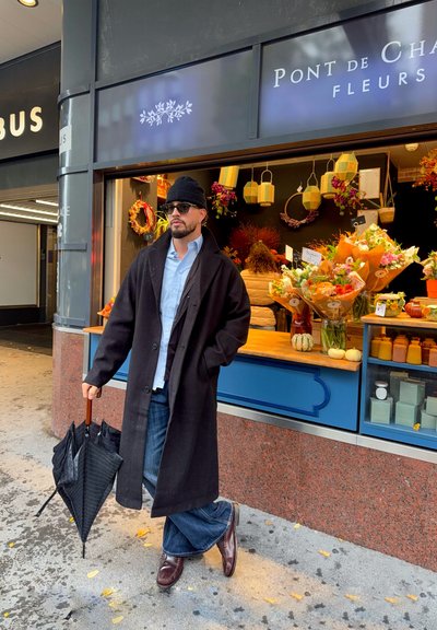 Abrigo negro, camisa azul y jeans acampanados. Sosteniendo un paraguas negro con un mango de madera. Tienda de flores con coloridos ramos en el fondo.