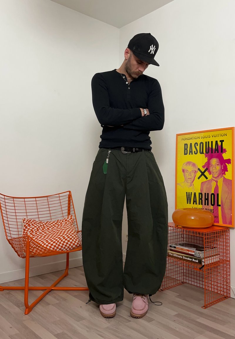 Homme en chemise noire à manches longues et pantalon large vert, debout les bras croisés à côté d'une chaise en fil orange et d'une affiche d'art Basquiat-Warhol.