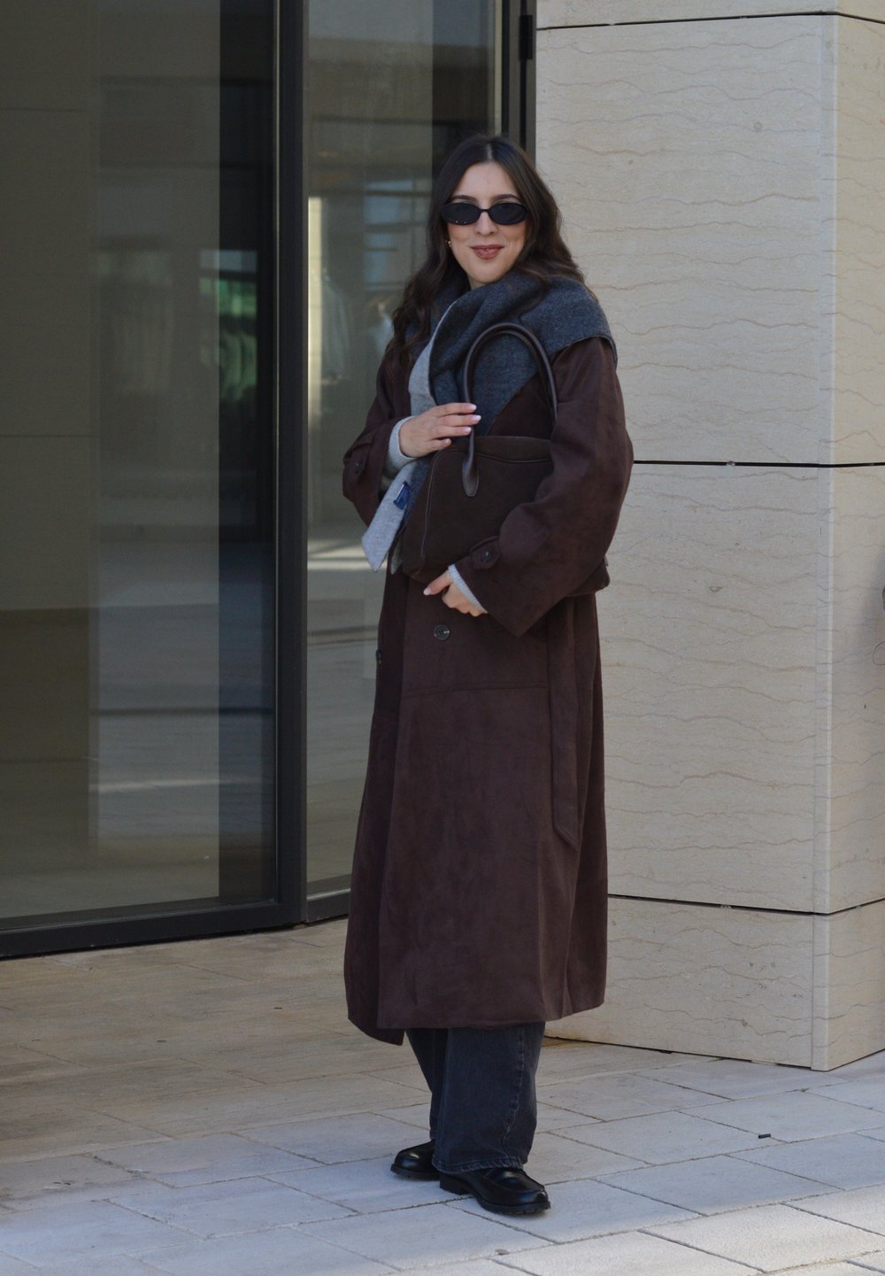 Long manteau marron en daim avec de gros boutons, écharpe grise et sac à main noir. Porté avec un jean foncé et des chaussures. Porte en verre transparent en arrière-plan.