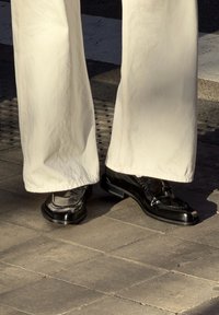 Personne portant un pantalon blanc à jambes larges et des mocassins en cuir noir brillant, debout sur un pavement carrelé au soleil.