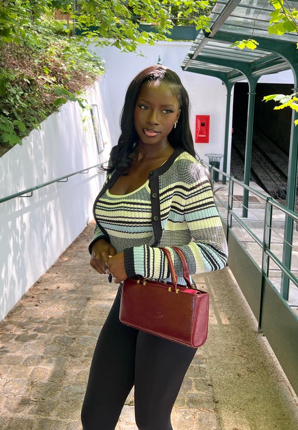 Jeune femme portant un pull rayé et un pantalon noir se tient sur un chemin pavé ensoleillé, tenant un sac à main rouge près de l'entrée d'un escalier extérieur.