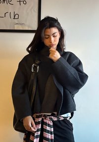 Jeune femme aux cheveux foncés portant une veste sombre surdimensionnée, un haut noir et une chemise rayée nouée à la taille, ajustant son col à l'intérieur.
