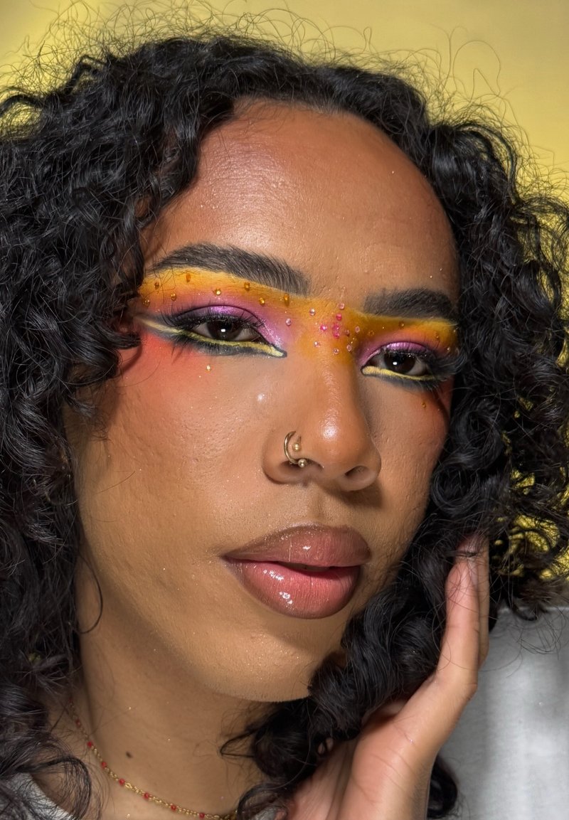 Person mit lockigem Haar, lebhaftem gelb-rosa Augen-Make-up mit kleinen Perlen und einem goldenen Nasenring, Hand auf Gesicht auf gelbem Hintergrund ruhend.