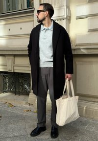 Vīrietis melnā mēteli, pelēkā džemperī un biksēs tur bežīgu iepirkuma maisiņu, stāv ārā uz akmens bruģa pie grezniem ēkas fasādes.