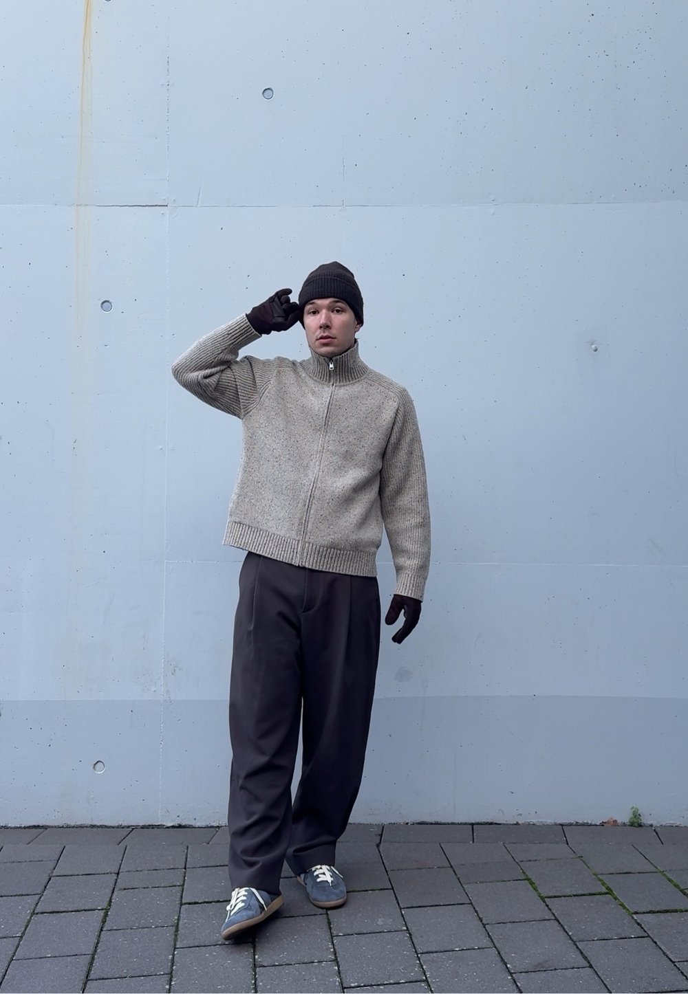 Pull beige en tricot avec fermeture éclair et col montant, associé à un pantalon ample foncé et un bonnet marron, debout sur une surface en pierre.