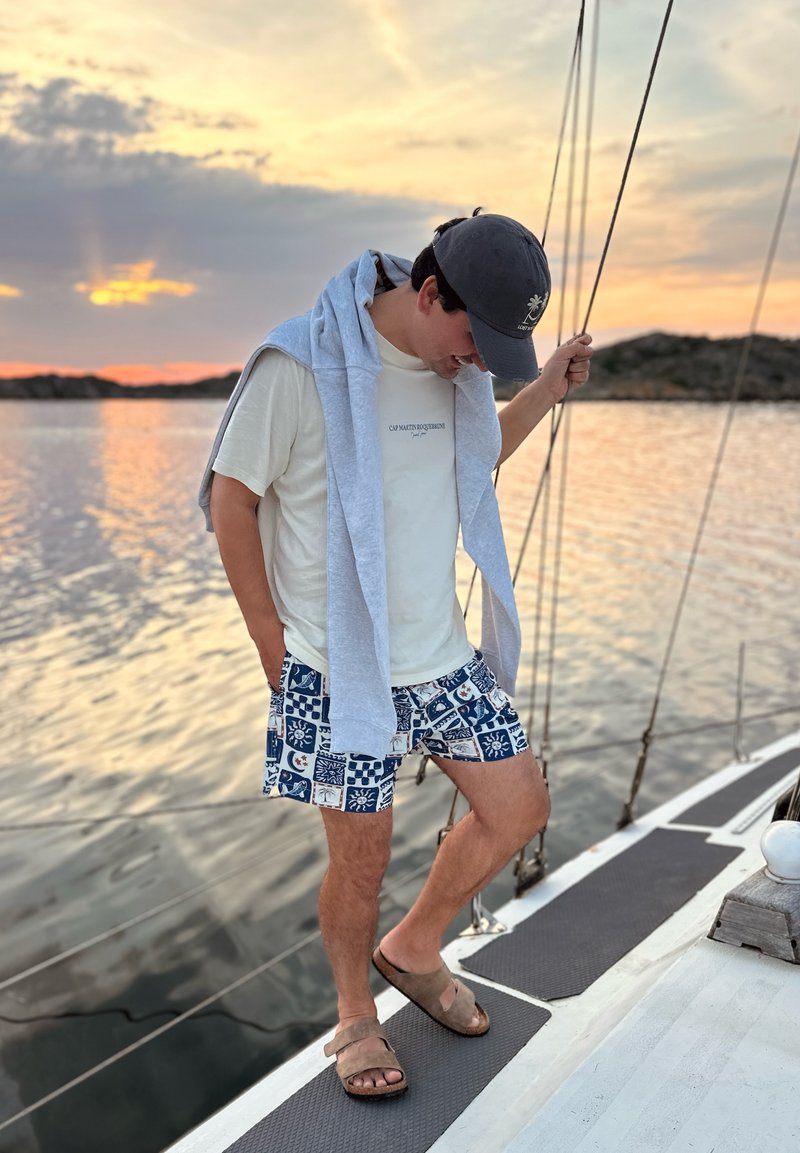 Homme debout sur un bateau au coucher du soleil, portant une casquette, un t-shirt blanc, un short à motifs, des sandales et un sweat-shirt gris posé sur ses épaules.