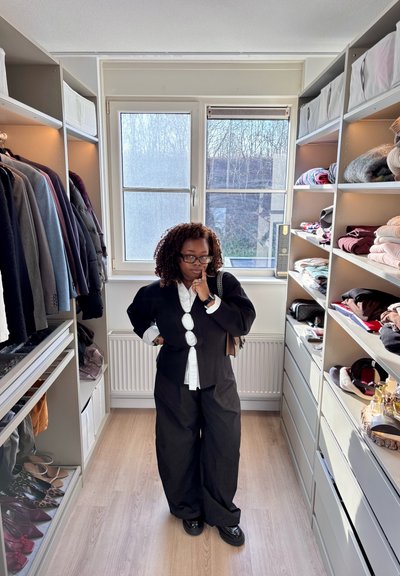 Mujer con atuendo blanco y negro de pie pensativa en un vestidor bien organizado con ropa y accesorios en estantes.