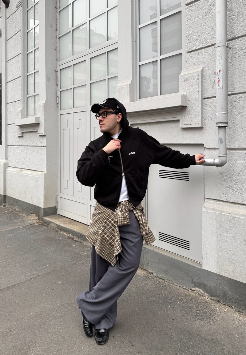 Man in black cap, glasses, black zip jacket, plaid shirt tied at waist, grey trousers, leaning on white building pipe on urban street.