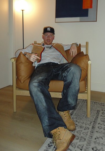 Silla de madera con grandes cojines marrones; figura con suéter gris y chaleco beige; lleva jeans desgastados y botas color marrón claro; suelo de madera clara.