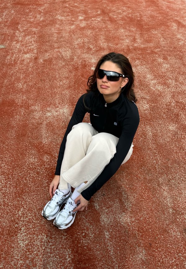 Femme portant des lunettes de soleil, une veste noire et un pantalon crème, assise sur une piste de sport rouge, genoux relevés, mains tenant ses baskets blanches.