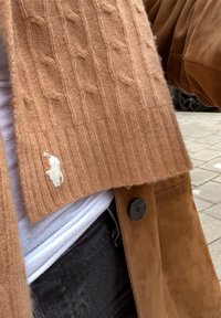 Beżowy sweter dzianinowy z prążkowanym dołem i małym, białym haftowanym logo. Noszony na białej koszuli i czarnych dżinsach.