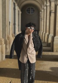 Palton lung texturat negru, sacou bej și pantaloni negri cu paiete. Ochelari de soare reflectanți. Plimbându-se într-un culoar cu coloane și podea din piatră.