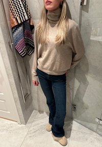 Μπεζ πλεχτό ζιβάγκο, μπλε φαρδιά τζιν και ανοιχτό καφέ παντόφλες με γούνα. Πολύχρωμες πετσέτες με σχέδια κρέμονται στο παρασκήνιο.