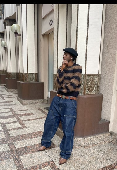 Suéter de punto con rayas marrones y negras, combinado con unos vaqueros de denim azul sueltos y zapatos de piel marrón. Los accesorios incluyen una boina negra y anillos.