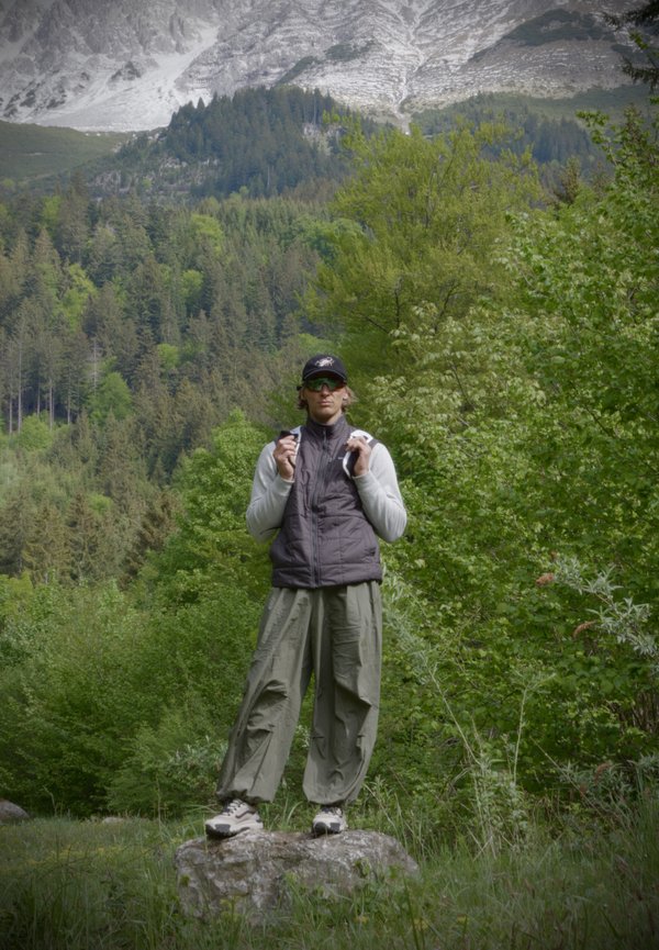 Person mit Sonnenbrille, Mütze und Outdoor-Bekleidung steht auf einem Felsen, umgeben von grünen Bäumen mit Wald und Bergen im Hintergrund.