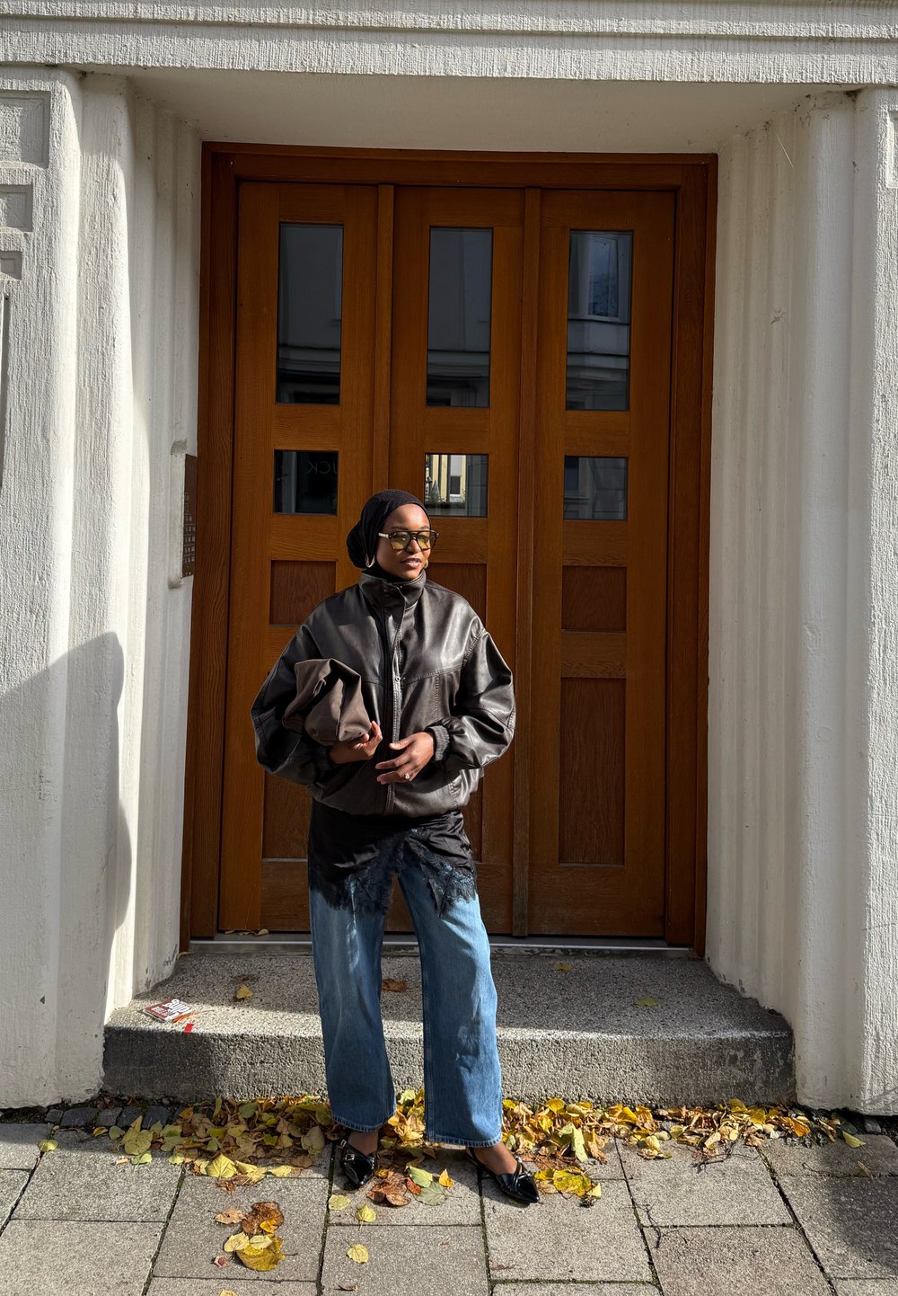 Crna kožna jakna, majica s čipkastim rubom, svijetloplave široke traperice i crne balerinke. Stoji ispred smeškastih dvostrukih vrata s četvrtastim prozorima.