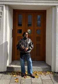 Crna kožna jakna, majica s čipkastim rubom, svijetloplave široke traperice i crne balerinke. Stoji ispred smeškastih dvostrukih vrata s četvrtastim prozorima.