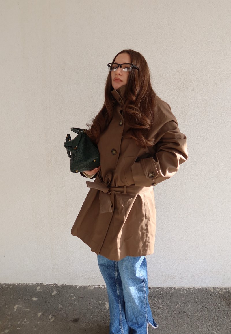 Donna con lunghi capelli castani che indossa occhiali, un cappotto marrone con cintura e jeans blu, che tiene una borsa verde scuro contro una parete semplice.