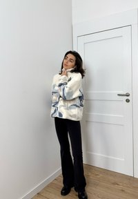 Løs hvid og blå fleecejakke med lynlås og høj krave, båret med sorte vide bukser og sorte ankelstøvler, sat op mod en ensfarvet væg.