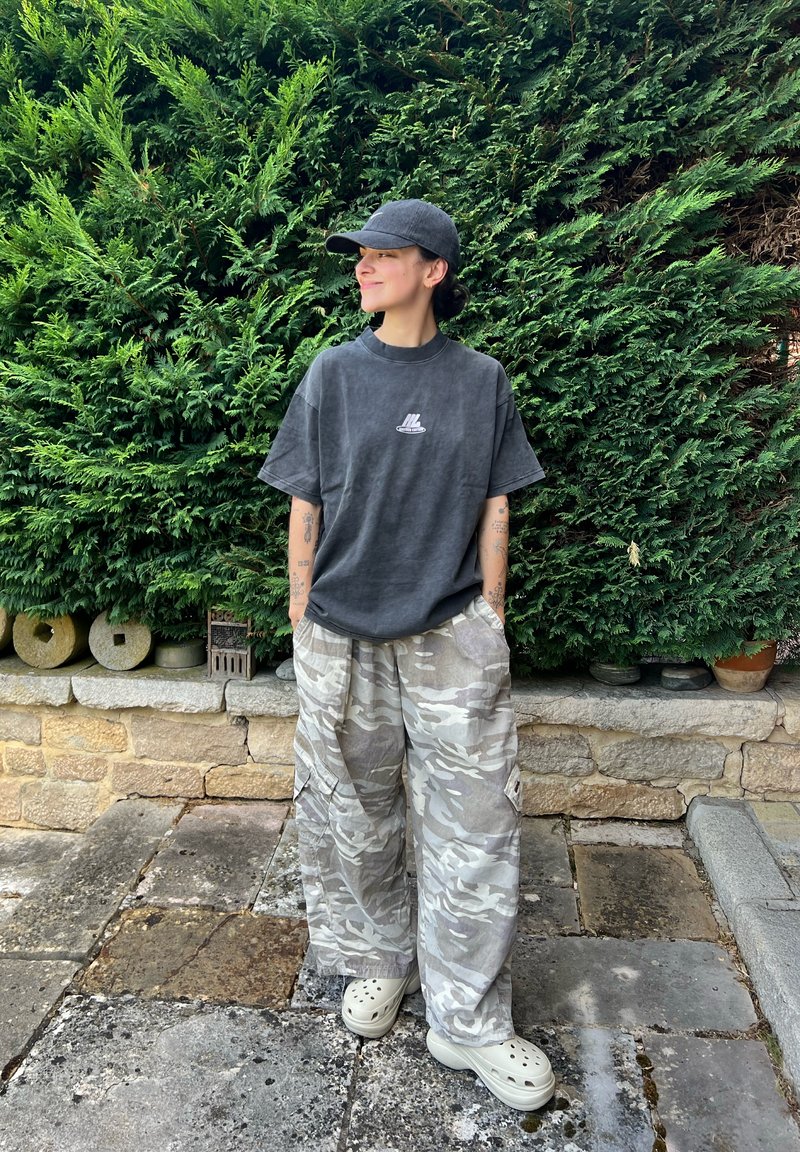 Personne portant une casquette sombre et une chemise avec un pantalon camouflage et des chaussures légères à enfiler, debout sur un pavement en pierre devant des buissons verts denses.