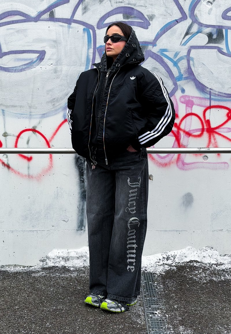 Personne portant une veste Adidas noire, un pantalon large noir avec le texte "Juicy Couture", des baskets à accents néon et des lunettes de soleil devant un mur de graffiti.