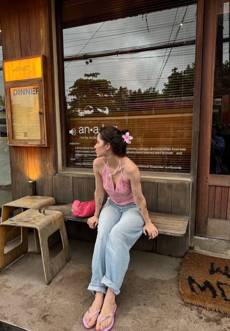 Kvinde med blomst i håret, lyserød top, lyse jeans og flip-flops, siddende på en træbænk udenfor en restaurant med en middagsmenu vist.