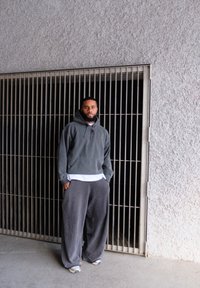 Pilkas džemperis su kapisonu, dėvimas ant baltos marškinėlių, derinamas su laisvais pilkais sportiniais džinsais ir šviesiais sportbačiais, prieš tekstūruotą sieną.