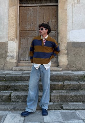 Hombre con cabello rizado y gafas de sol de pie en unas escaleras de piedra, lleva un suéter a rayas marrón y azul sobre una camisa blanca y unos jeans anchos de color azul claro.