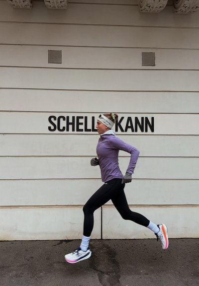Mujer con camiseta morada de manga larga y leggings negros corriendo frente a una pared blanca con texto negro que dice "SCHELL KANN."