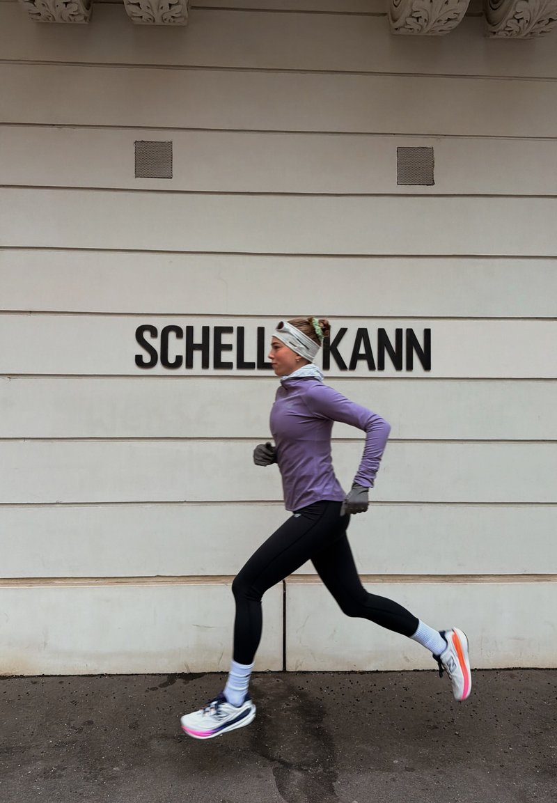 Mujer con camiseta morada de manga larga y leggings negros corriendo frente a una pared blanca con texto negro que dice "SCHELL KANN."