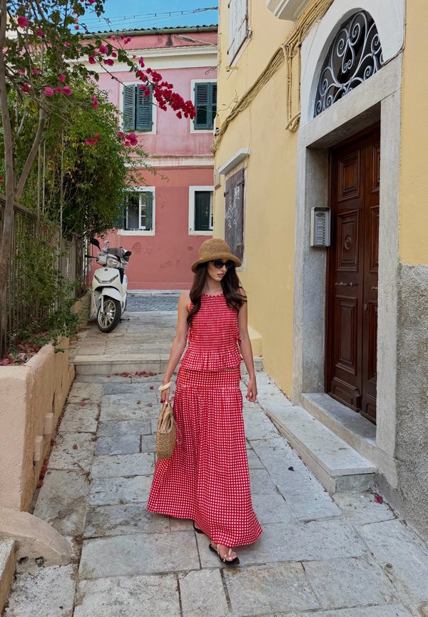 Femme en robe rouge à carreaux et chapeau de soleil marchant sur un chemin en pierre passant devant des bâtiments colorés et un scooter blanc garé près d’un mur rose.