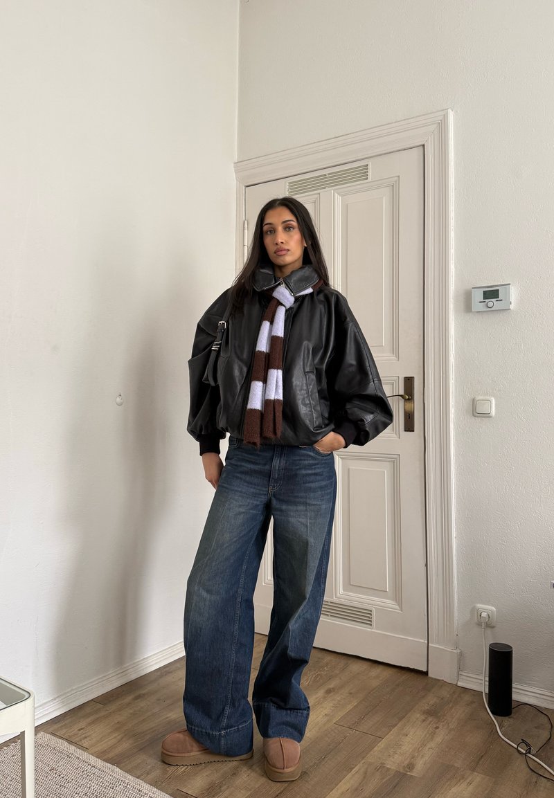 Femme debout à l'intérieur portant une veste en cuir noire, une écharpe rayée, un jean à jambes larges et des chaussures compensées couleur caramel, avec une main dans la poche.