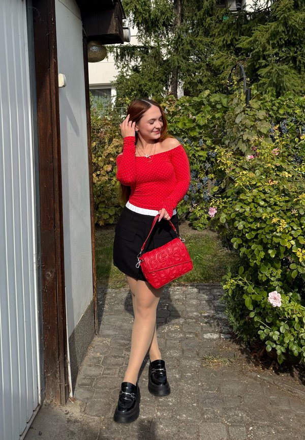 Mujer con camiseta roja de manga larga y falda negra está de pie en un camino de piedra cerca de la vegetación, tocándose el cabello y sosteniendo un bolso rojo.