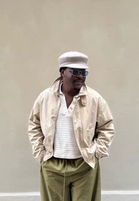 Beige bomberjack, wit gebreid shirt, olijfgroene broek, lichtgekleurde pet, ronde zonnebril, staand tegen een neutrale achtergrond.