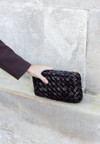 Hand die een zwart geweven leren clutch vasthoudt tegen een textuur van een lichte stenen muur als achtergrond.