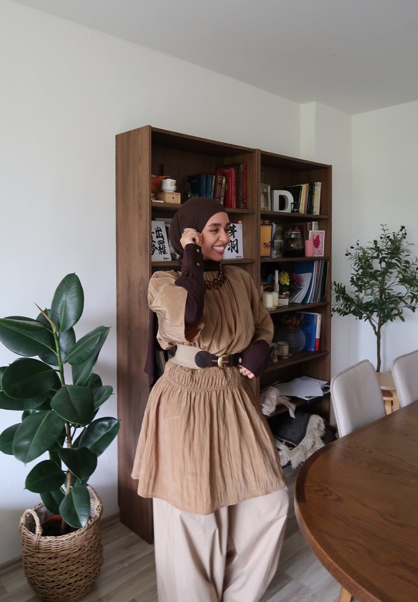 Nő barna hijabot és bézs ruhát visel, mosolyog egy fa könyvespolc mellett, amely könyvekkel és növényekkel teli, egy világos nappaliban.