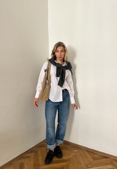 Camisa blanca de botones, bufanda gris oscuro atada al cuello, bolso beige, jeans azul claro y zapatos negros, de pie contra una pared clara en una esquina.