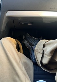 Schwarze Sportschuhe aus strukturiertem Material mit reflektierenden Akzenten, getragen zu beigen Hosen, im Inneren eines Fahrzeugs.