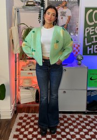 Femme debout à l'intérieur portant un cardigan vert à motifs, un crop top blanc, un jean évasé foncé et des chaussures noires, les mains dans les poches.