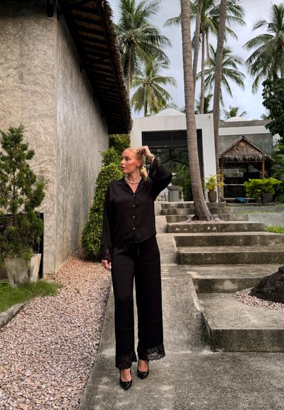 Blusa negra de satén de manga larga con puños de encaje, combinada con pantalones de pierna ancha a juego y tacones altos negros. Entorno al aire libre con vegetación.