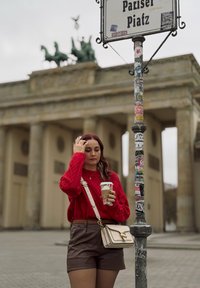 Suéter de punto rojo, pantalones cortos de cuero marrón y bolso beige. Sosteniendo una taza de café. La Puerta de Brandeburgo de Berlín de fondo con un cartel.