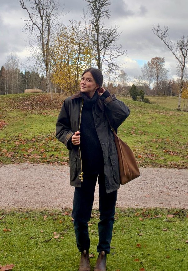 Olivově zelená nepromokavá bunda se zapínáním na zip, černý rolák, černé kalhoty a hnědá kožená kabelka. Přírodní prostředí s stromy.