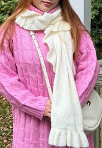 Γυναίκα που φοράει ροζ πουλόβερ πλεκτό με κοτσίδες και λευκό ριγωτό φουλάρι, κρατώντας μια μπεζ τσάντα ώμου σε εξωτερικό χώρο κοντά σε φυτά.