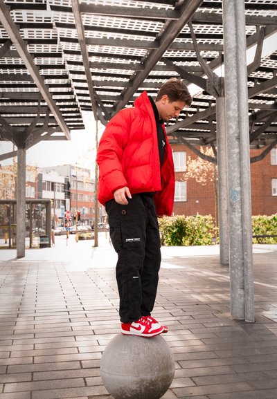 Chaqueta de abrigo roja sobre una camiseta negra, combinada con pantalones cargo negros y zapatillas rojas y blancas, de pie sobre una esfera de concreto al aire libre.
