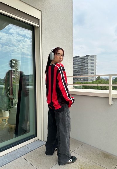 Camisa de manga larga a rayas rojas y negras, pantalones anchos negros y zapatillas verdes. Se lleva grandes auriculares blancos, de pie en un balcón.