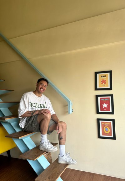 Escalera de madera con pasamanos azul; individuo con camisa blanca exagerada y pantalones cortos grises; carteles de viaje enmarcados en la pared; fondo claro.
