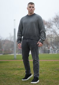 Pull gris côtelé avec un col noir, jean gris foncé et baskets noires à semelles blanches, posés devant un fond herbeux.